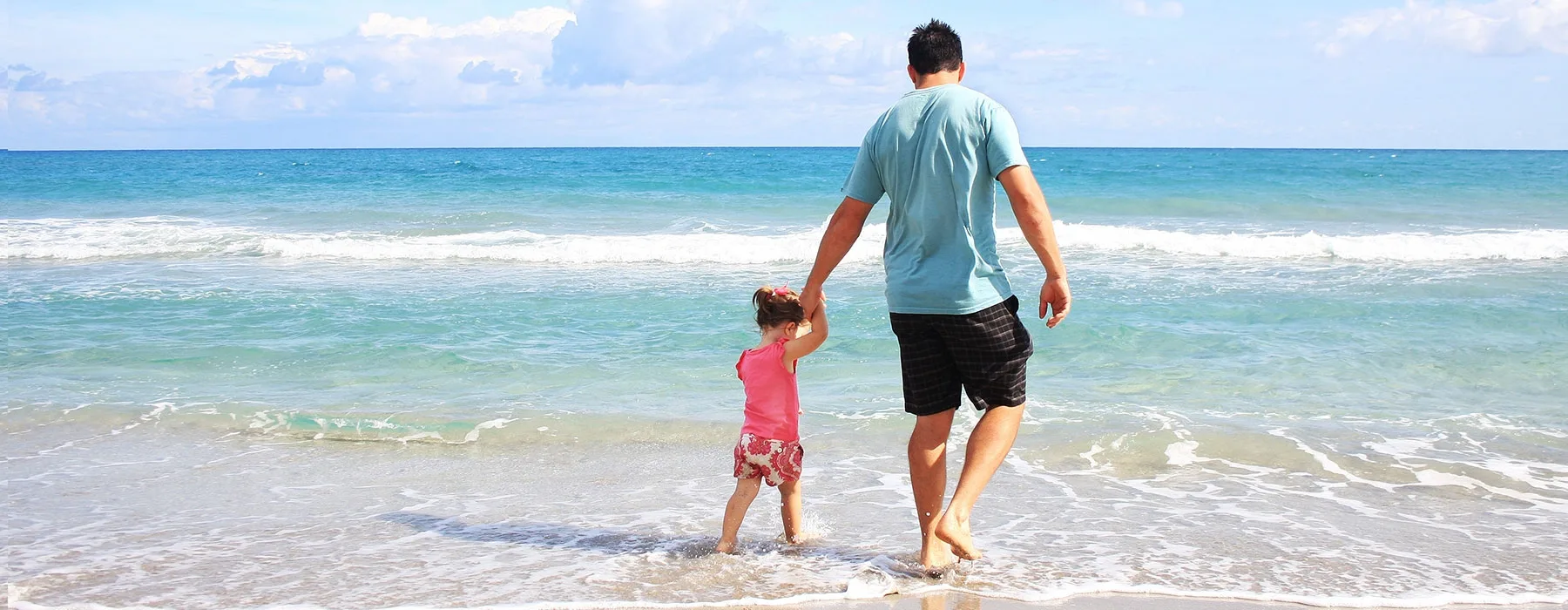 father and young daughter holding hands while wading into the water on the beach